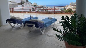 Un balcón con dos tumbonas, una planta en maceta y vistas al mar y edificios.