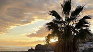 A swimming pool with a palm tree in the foreground, a sunset in the background, and buildings across the water. - Playa del Sol – Sea View Studio in Puerto Rico (Mogán)