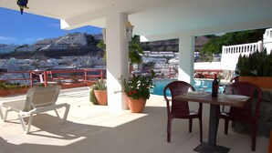 A covered outdoor area with a table and chairs, potted plants, and a view of a coastal town. - Playa del Sol – Sea View Studio in Puerto Rico (Mogán)