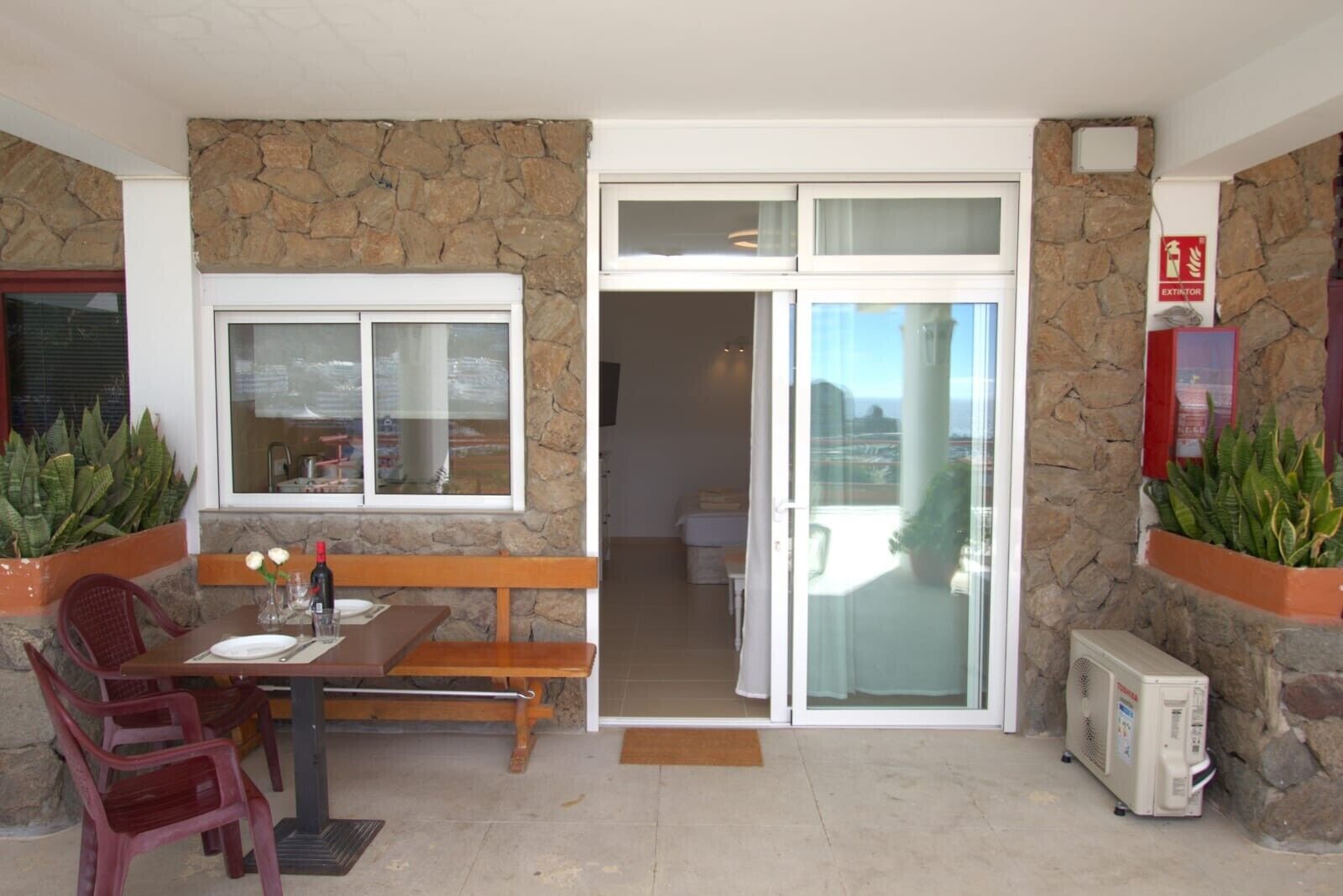 Uma área de pátio com mesa, cadeiras, banco, geladeira e plantas em vasos.