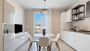 A modern kitchen with white cabinetry, a dining table with chairs, a pendant light, and a view through the window.