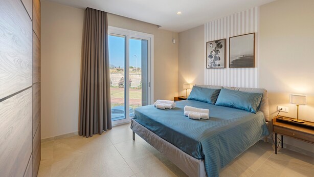 A bedroom with a bed, a nightstand, a lamp, a window with curtains, and a view of the outside.