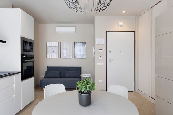 A modern kitchen with built-in appliances, a dining table with a potted plant, a grey sofa, and a white door.