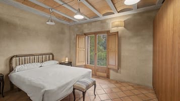 Un dormitorio con cama, mesita de noche, banco y ventana con contraventanas.