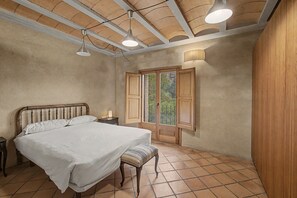 Quarto com cama, mesa de cabeceira, banqueta e janela com portaria.
