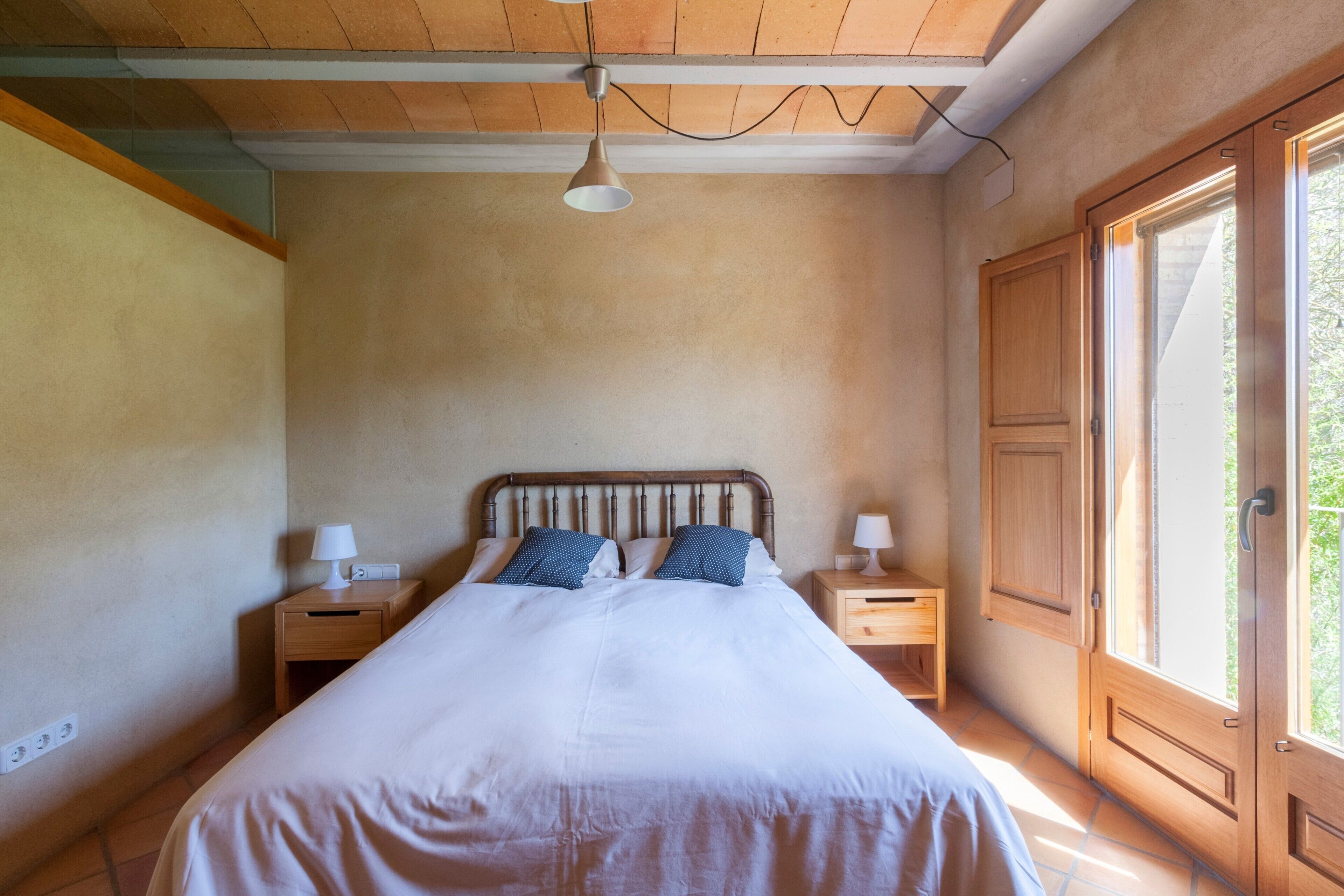 A bedroom with a wooden ceiling, a bed with white bedding, two nightstands with lamps, and a window with a view of greenery.