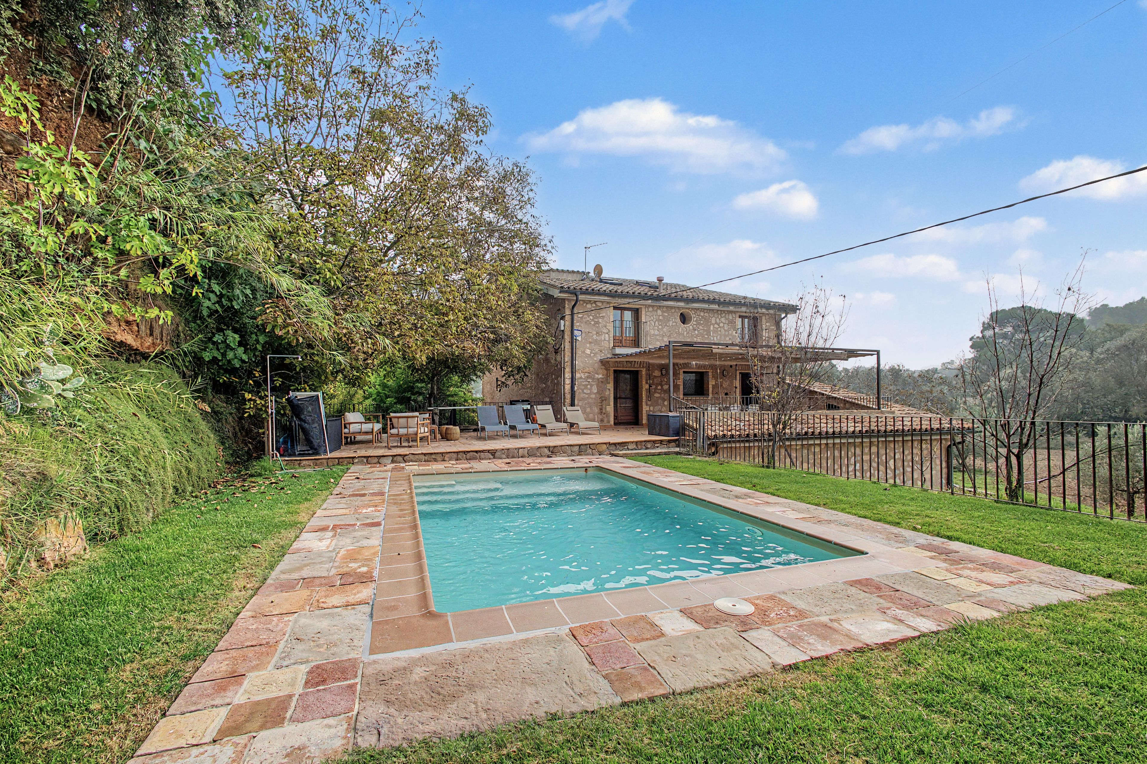 Una piscina con borde de piedra, un patio con sillas y una casa de piedra con techo de tejas.