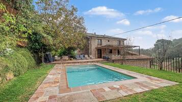 Una piscina con borde de piedra, un patio con sillas y una casa de piedra con techo de tejas.