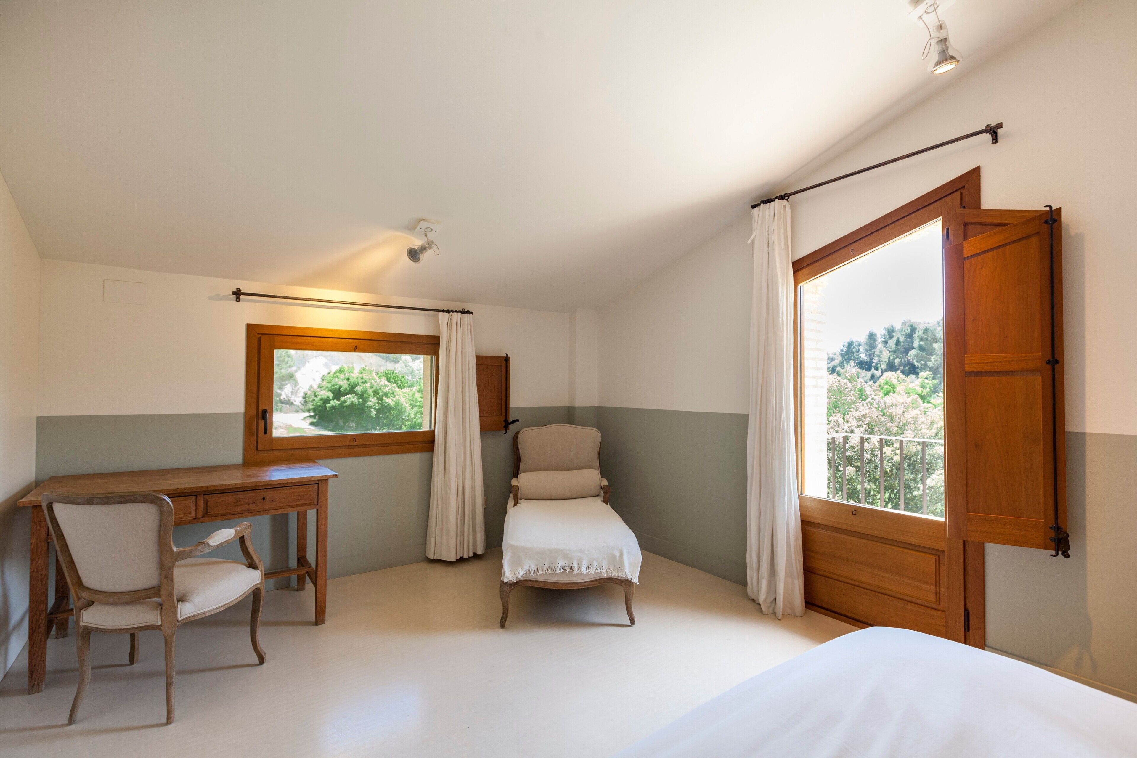 Un dormitorio con cama, escritorio, silla, ventana con cortinas y puerta al balcón.