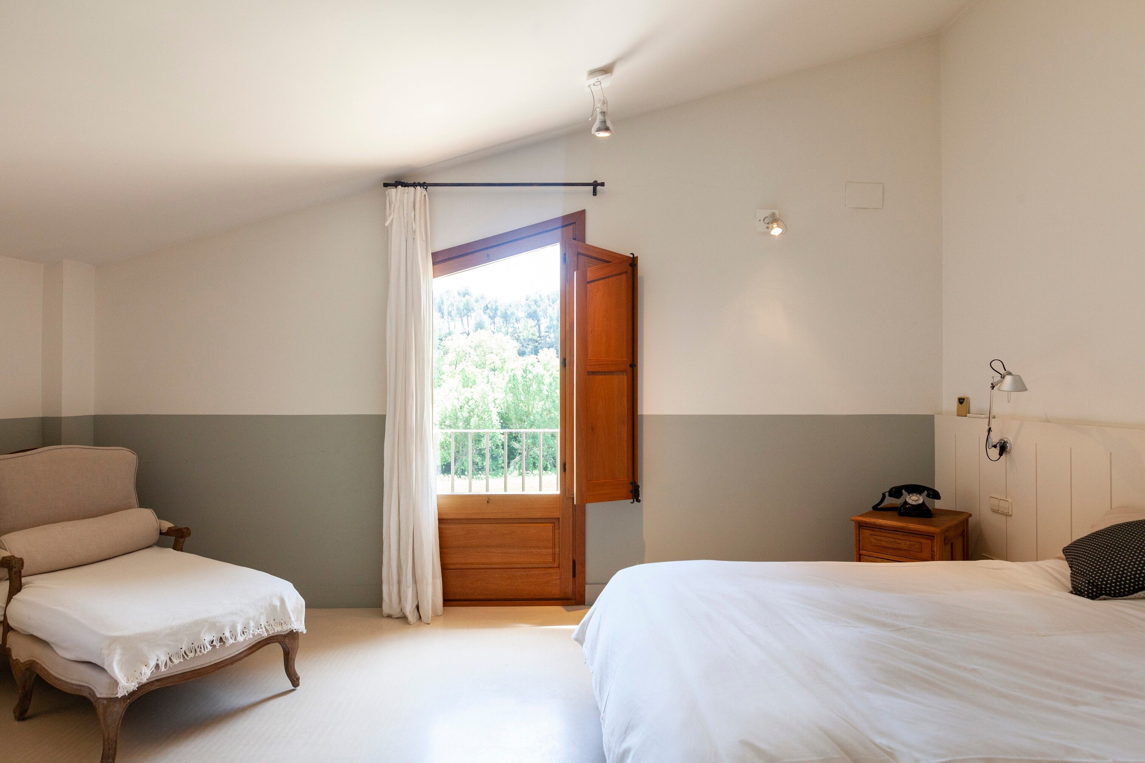 Un dormitorio con cama, una silla, una mesita de noche, una lámpara y una ventana con cortinas.