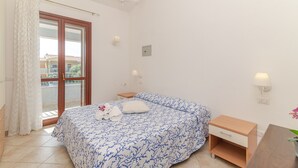 Quarto com cama, mesa de cabeceira, escrivaninha, candelabro, janela com cortinas e vista para prédios.