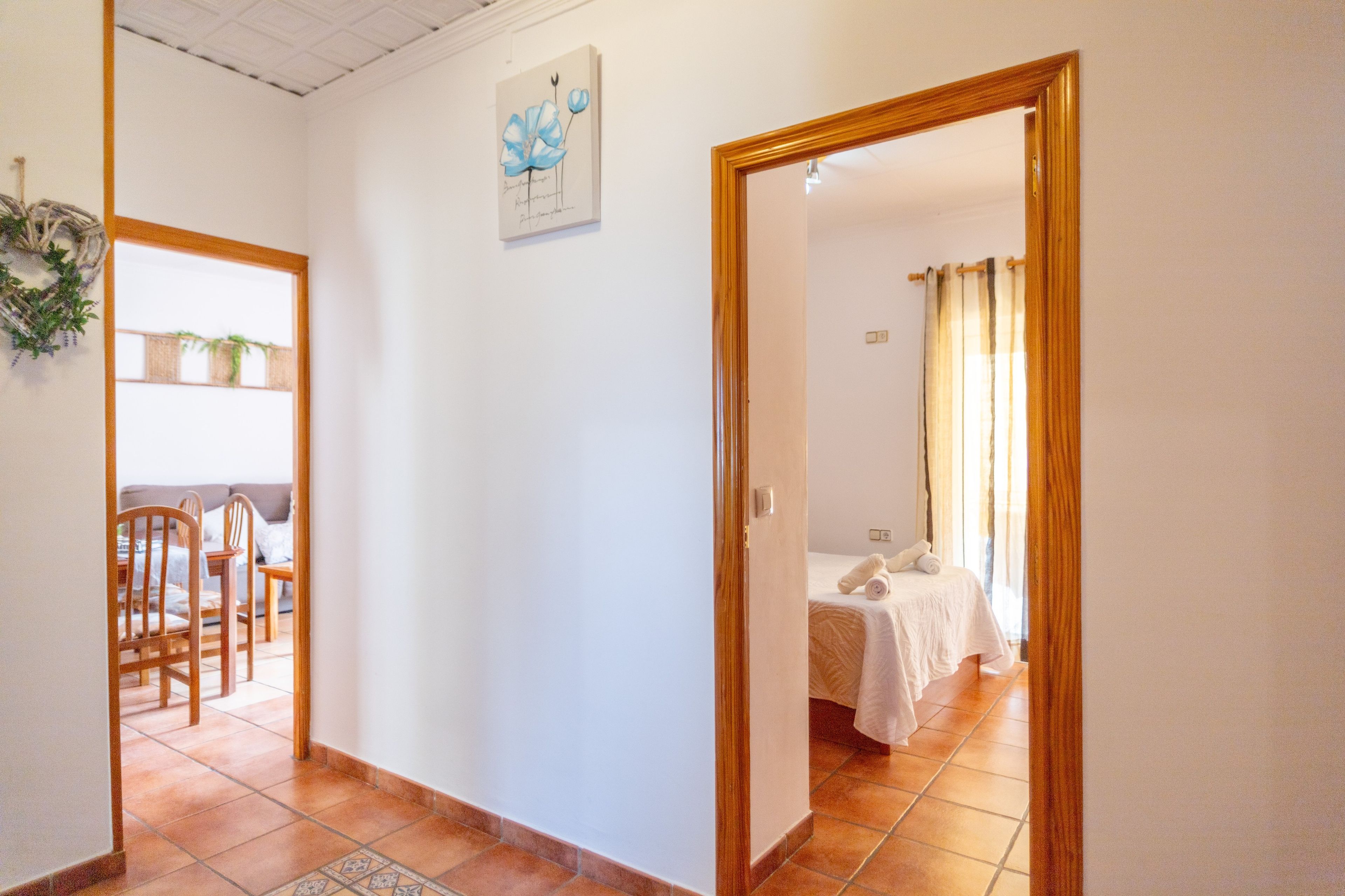 A hallway leading to a bedroom with a bed, a dining area with chairs, and a wall-mounted artwork.