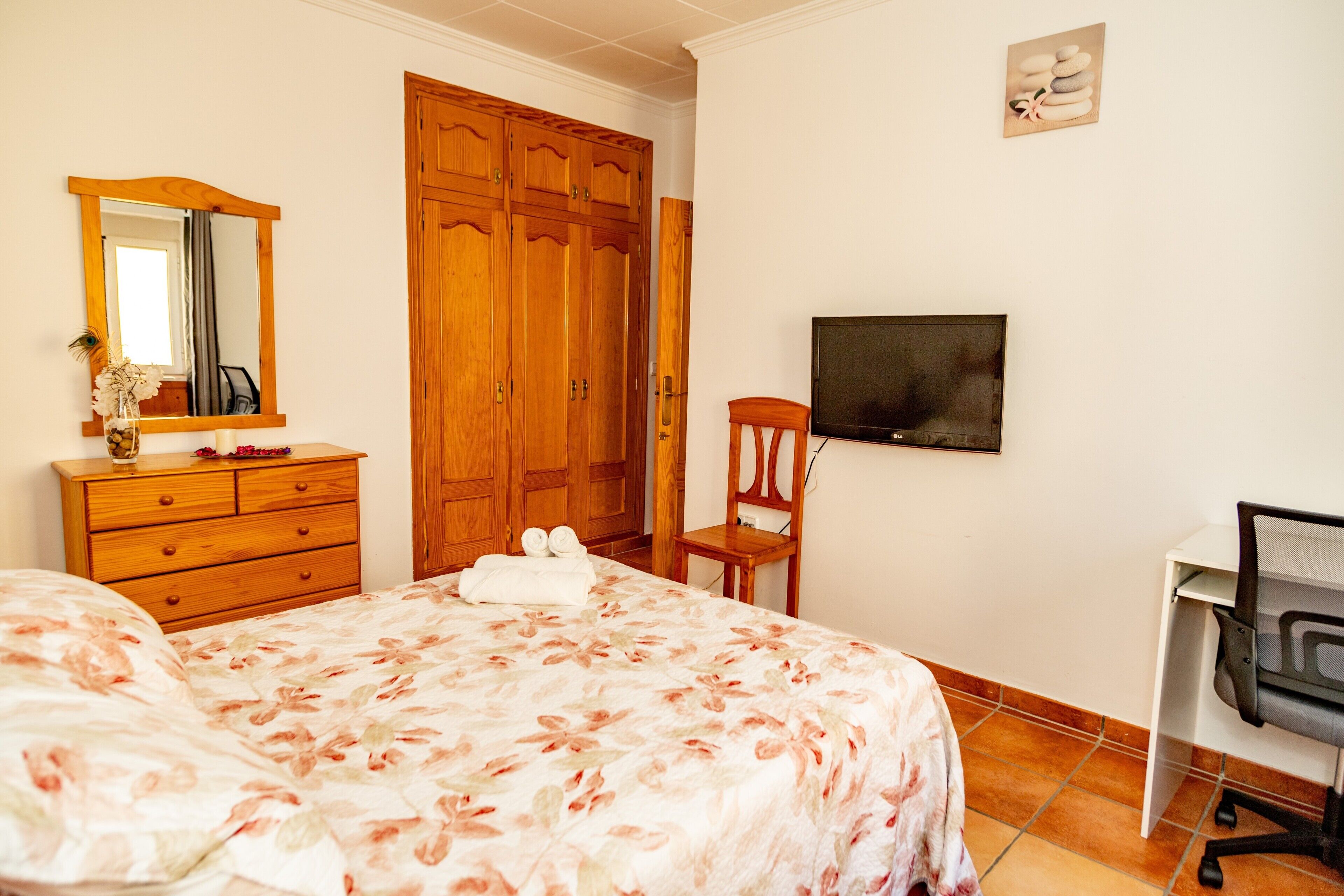 A bedroom with a bed, wooden wardrobe, a desk with a chair, a television, and a mirror.