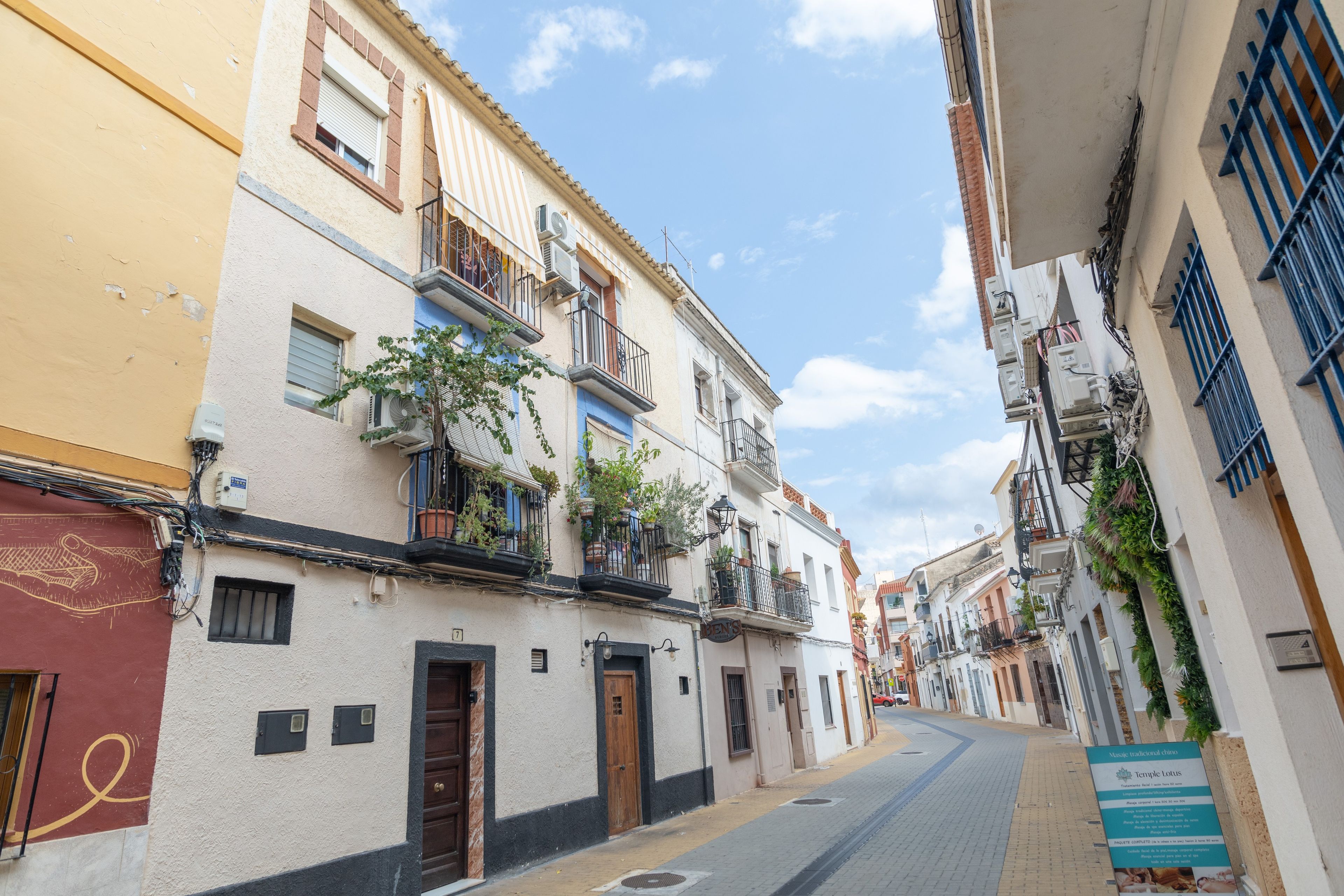 A narrow street lined with traditional buildings, some with balconies and potted plants.