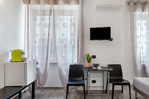 Una camera piccola e moderna con un letto, una scrivania con sedia, una pianta in vaso e una TV a parete.