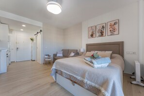 Una camera da letto moderna con un letto grande, un comodino, una piccola scrivania e un divano. Si vede una porta che conduce in un'altra stanza e una lampada a parete.
