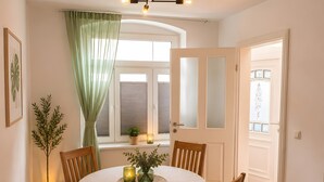 Zona pranzo con tavolo rotondo, sedie di legno, lampadario, pianta in vaso e finestra con tende.