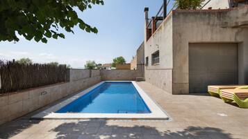 Una piscina rettangolare con bordo piastrellato, un cancello chiuso del garage e una pila di sedie a sdraio.