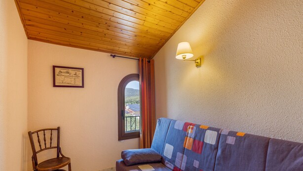 Une pièce avec un plafond en bois, une fenêtre avec des rideaux, un tableau encadré au mur, une chaise en bois et un canapé recouvert d’une couverture colorée.