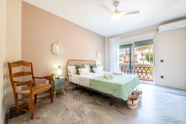 Ein Schlafzimmer mit Bett, Stuhl, Nachttisch, Deckenventilator und Blick ins Freie.