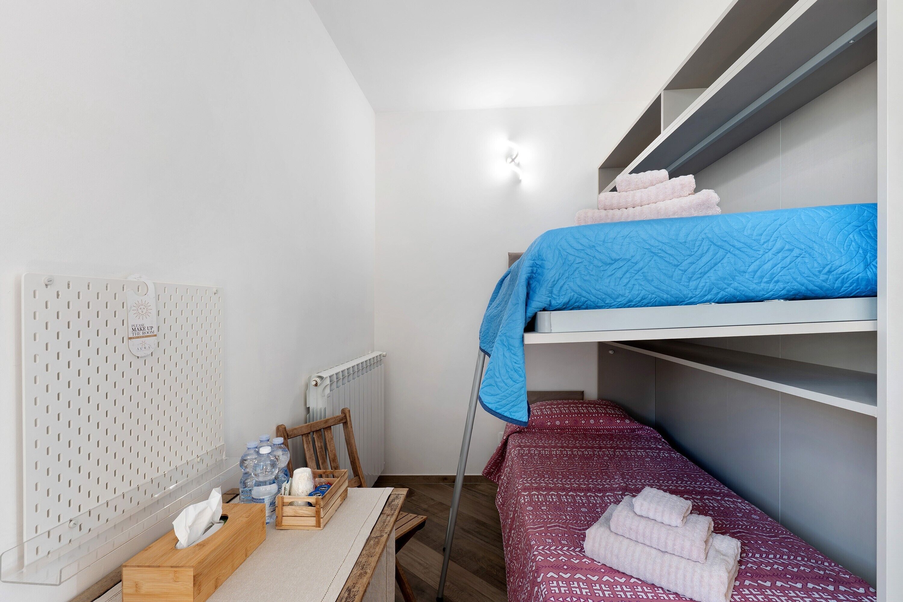A dormitory room with bunk beds, a radiator, a pegboard, a wooden chair, and a table with a tissue box and water bottles.