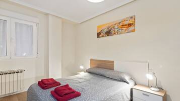 Un dormitorio con cama, dos toallas rojas, una mesita de noche con lámpara, un radiador y un cuadro en la pared.