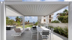 Un patio exterior con una pérgola blanca, una larga mesa de comedor y varias sillas.