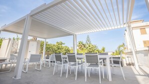 An outdoor patio with white chairs and tables, a pergola overhead, and a view of greenery and buildings in the background. - Villa 'Zefira Mare' with Sea View, Wi-Fi, and Air Conditioning (Altavilla Milicia)