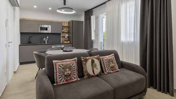 Una sala de estar moderna con un sofá gris, almohadones decorativos, una mesa de comedor y una cocina al fondo.