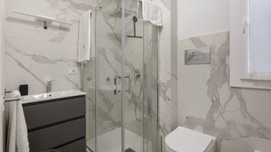 Un baño con ducha envuelta en vidrio, inodoro blanco, lavamanos con grifo y una toalla colgada en un perchero.