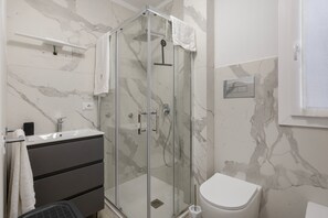 Ein Badezimmer mit einer ein Glasfach abgetrennten Dusche, einer weißen Toilette, einem Waschbecken mit Armatur und einem Handtuch an einem Haken.
