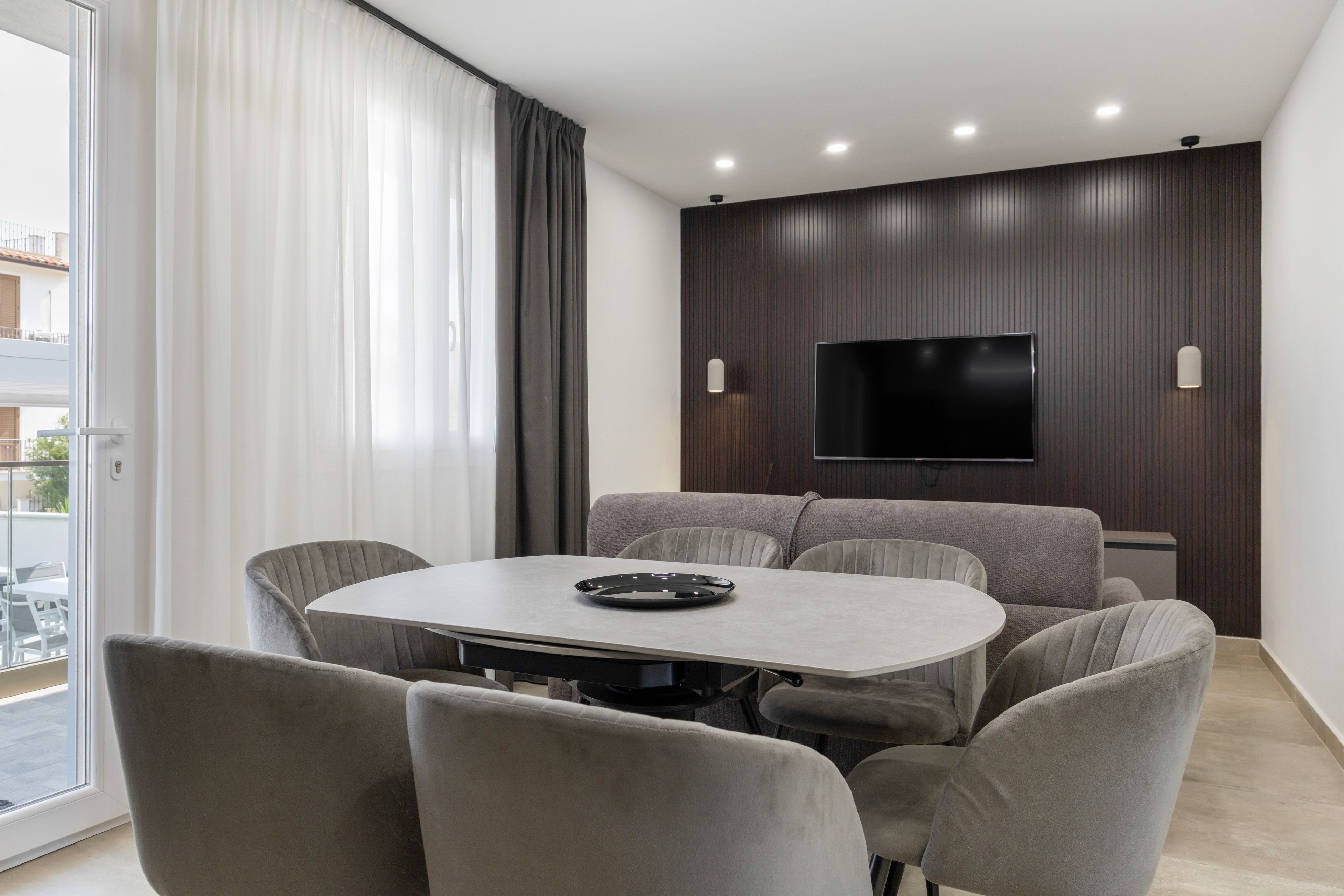 Una sala moderna con un televisor de pantalla plana montado en una pared de madera oscura, una mesa redonda de comedor con seis sillas grises tapizadas y un ventanal corredizo que da a un espacio exterior.