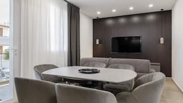 Una sala moderna con un televisor de pantalla plana montado en una pared de madera oscura, una mesa redonda de comedor con seis sillas grises tapizadas y un ventanal corredizo que da a un espacio exterior.