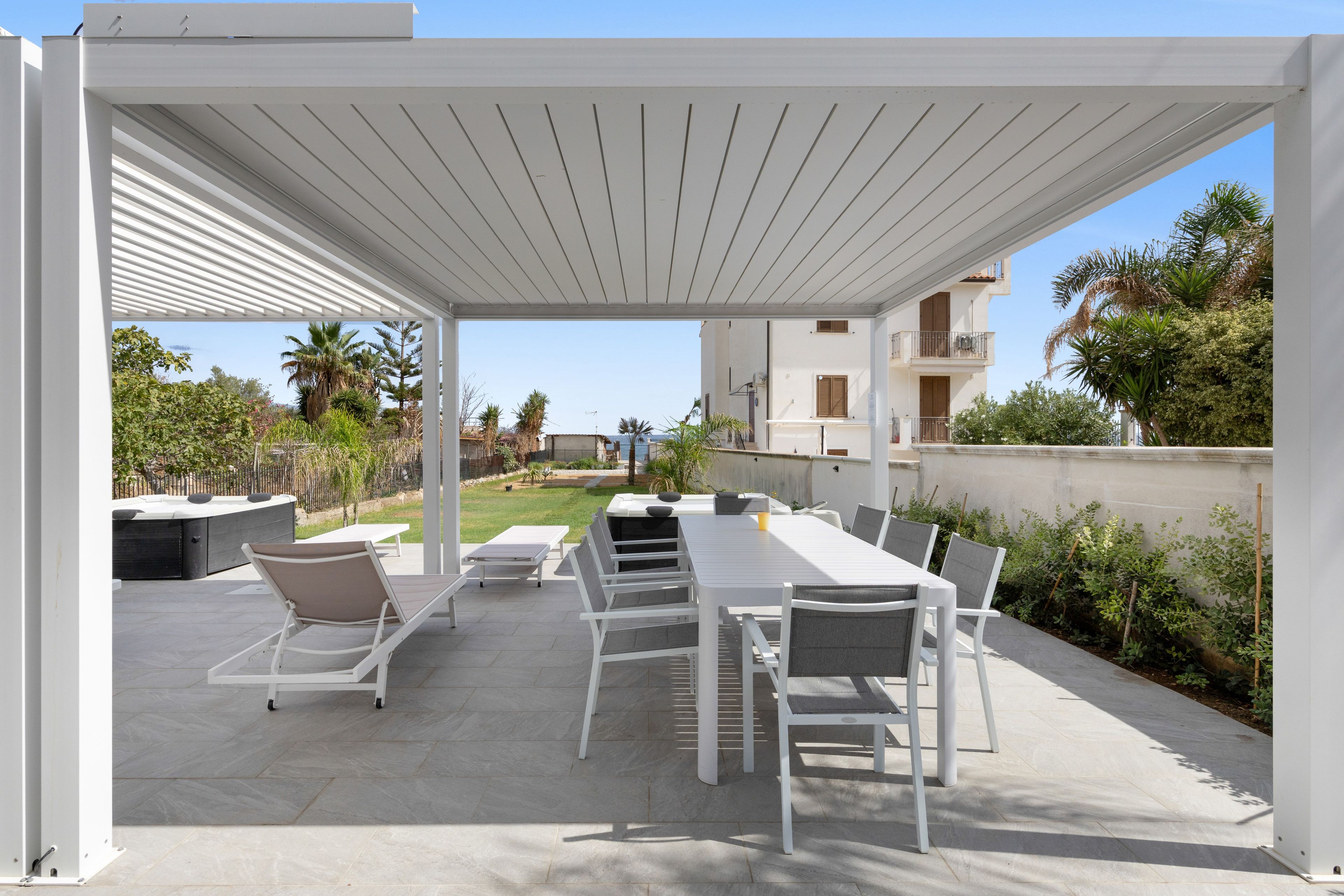 Un patio esterno con una pergola bianca, un lungo tavolo da pranzo con sedie e vista sul giardino.