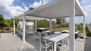 Un patio techado al aire libre con mobiliario blanco, que incluye una mesa, sillas y una parrilla.