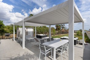 Eine überdachte Terrasse im Freien mit weißen Möbeln, darunter ein Tisch, Stühle und ein Grill.