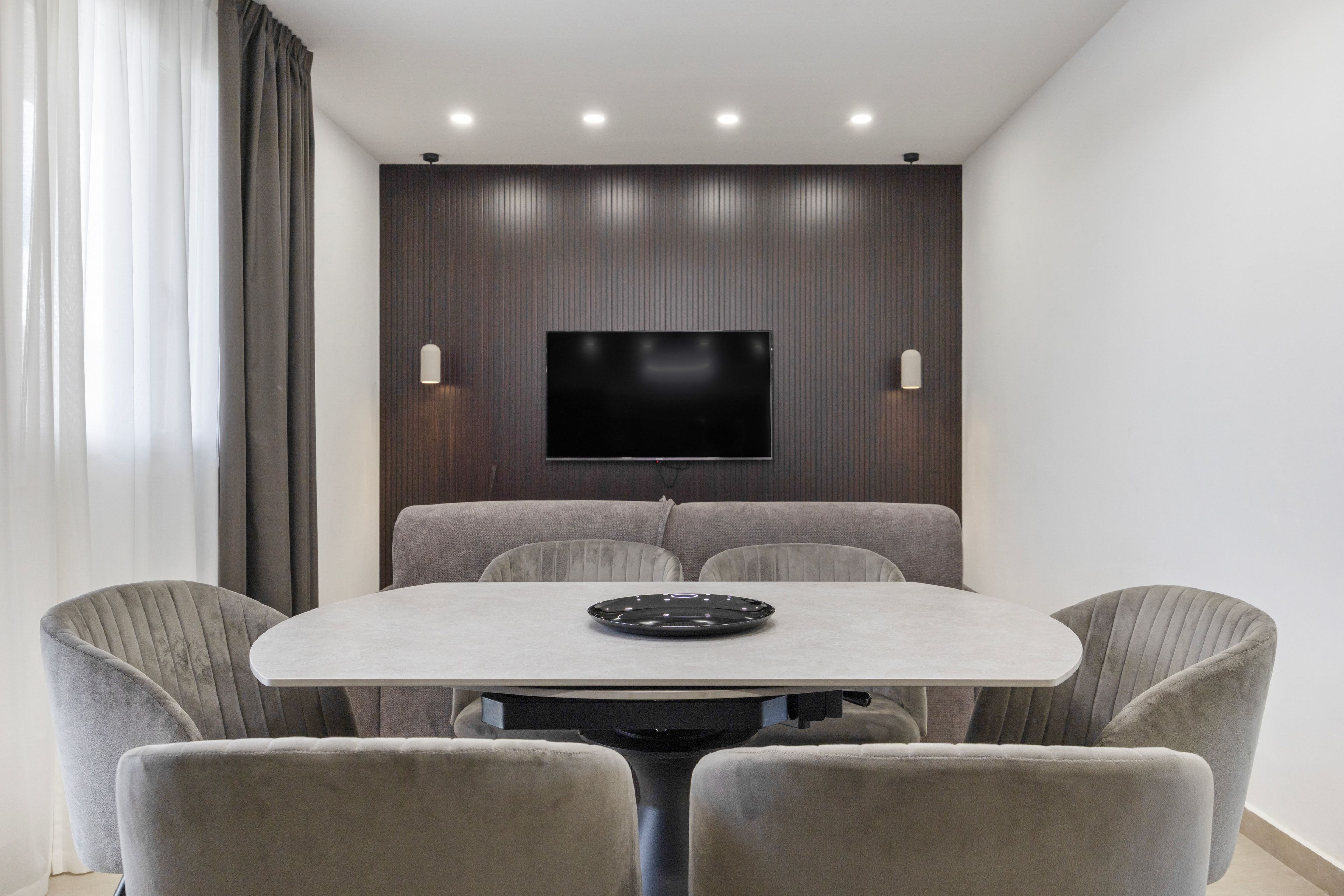 Un área de comedor moderna con una mesa redonda, cuatro sillas tapizadas, un televisor de pantalla plana montado en una pared de paneles de madera e iluminación empotrada en el techo.