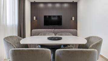 Un área de comedor moderna con una mesa redonda, cuatro sillas tapizadas, un televisor de pantalla plana montado en una pared de paneles de madera e iluminación empotrada en el techo.