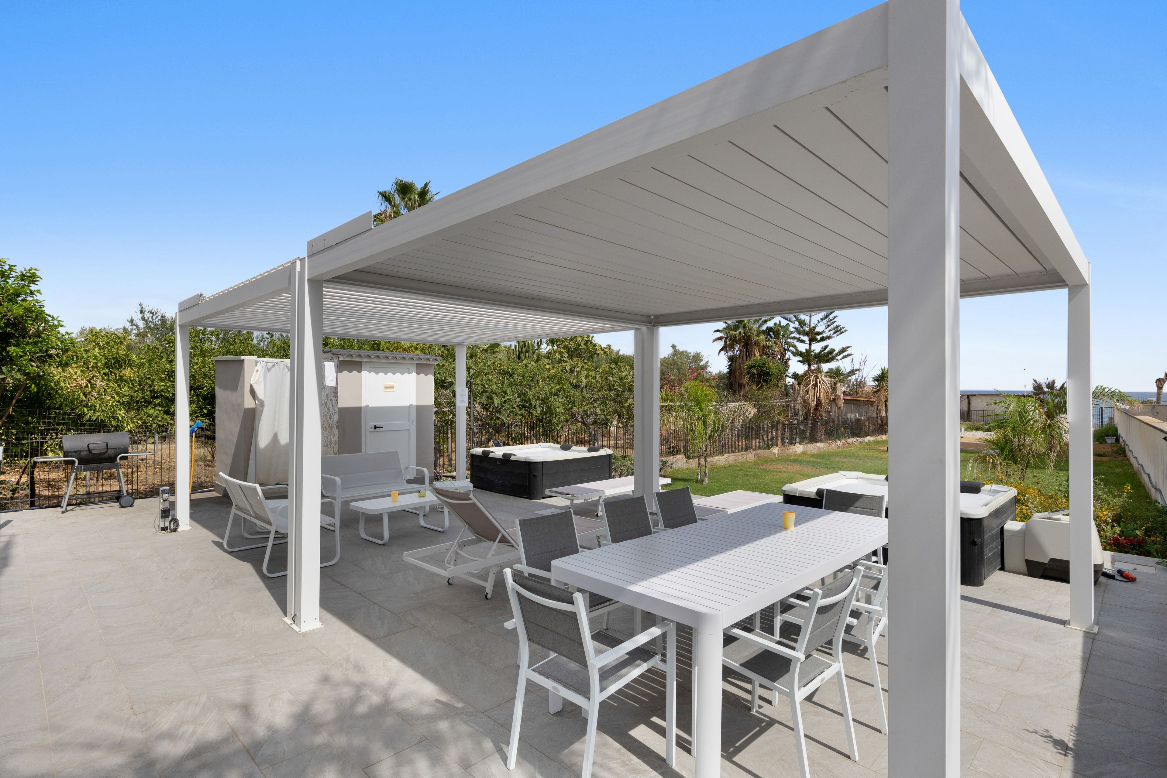 Un patio techado al aire libre con mobiliario blanco, incluyendo una mesa, sillas y una parrilla.