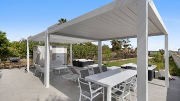 Un patio techado al aire libre con mobiliario blanco, incluyendo una mesa, sillas y una parrilla.