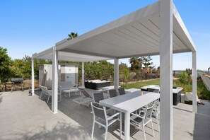 Een overdekt terras buiten met witte meubels, waaronder een tafel, stoelen en een barbecue.