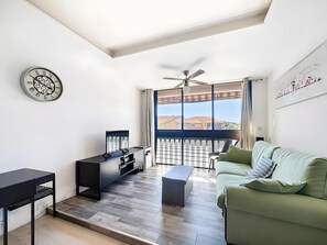 Un salon moderne doté d’un ventilateur de plafond, d’une grande fenêtre offrant une belle vue, d’un canapé vert et d’un meuble de rangement noir pour les appareils électroniques.