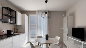 Une cuisine moderne avec des meubles blancs, une table à manger avec des chaises, un luminaire suspendu noir et un téléviseur posé sur un meuble blanc.