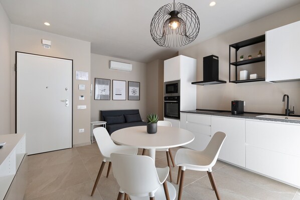 Une cuisine moderne avec des meubles blancs, une table de salle à manger ronde entourée de chaises, un luminaire suspendu noir et une petite plante en pot.