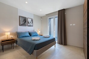 Une chambre à coucher comprenant un lit, des tables de chevet, une lampe, un tableau accroché au mur et une porte-fenêtre coulissante.