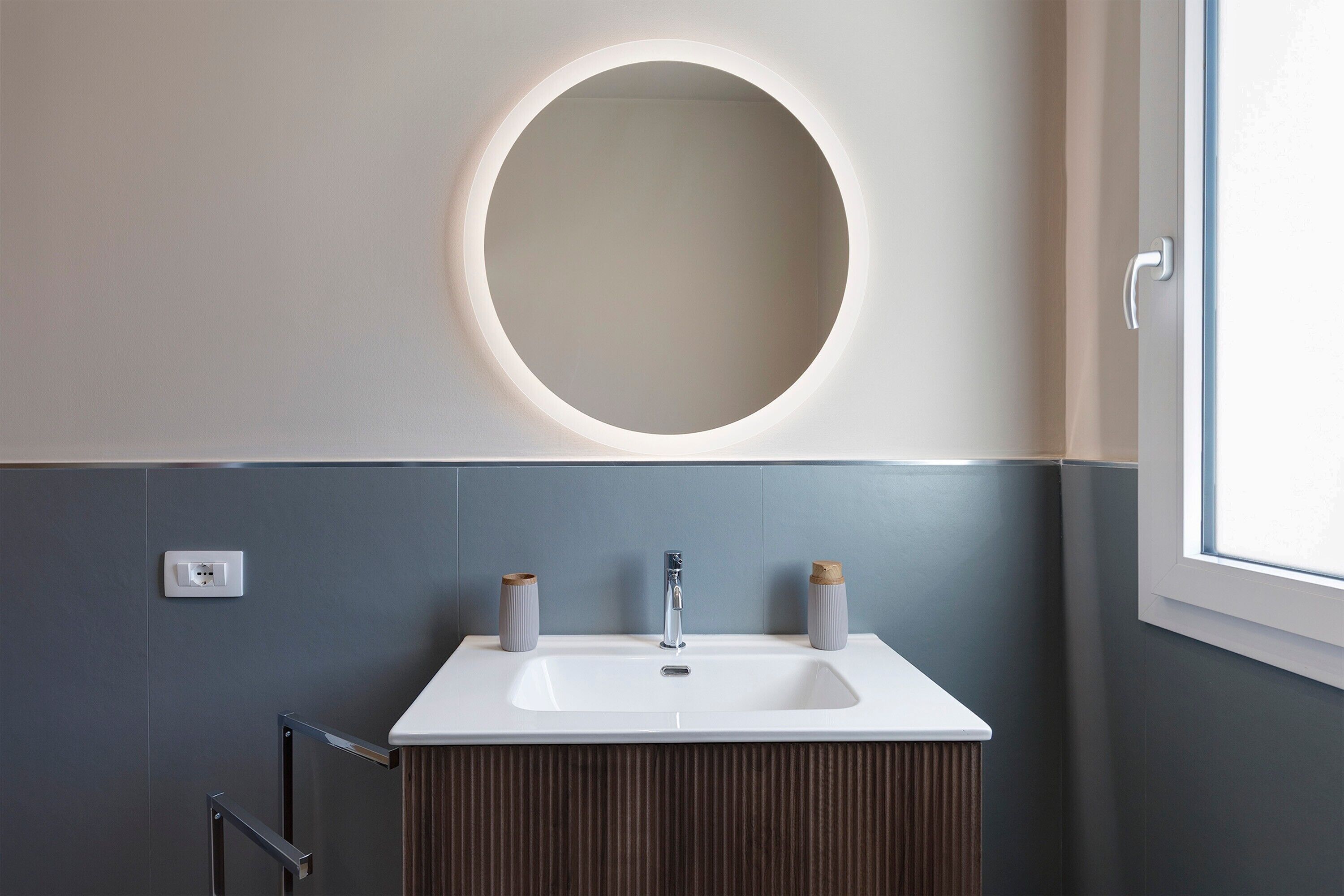 Une salle de bain avec un lavabo blanc, un miroir rond et deux récipients cylindriques posés sur le comptoir.