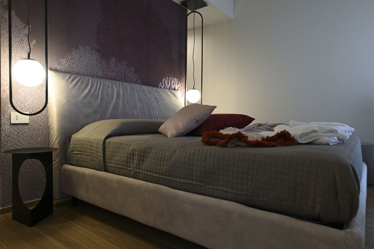 A modern bedroom with a large bed, two pendant lights, a small side table, and a textured wall.