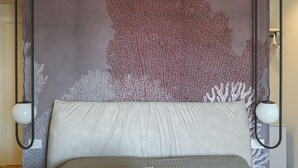 Sängyssä on harmaa patja ja valkoiset tyynyt, seinässä punavalkoinen korallikuvio ja lattiassa kaksi mustaa lattiavalaisinta.