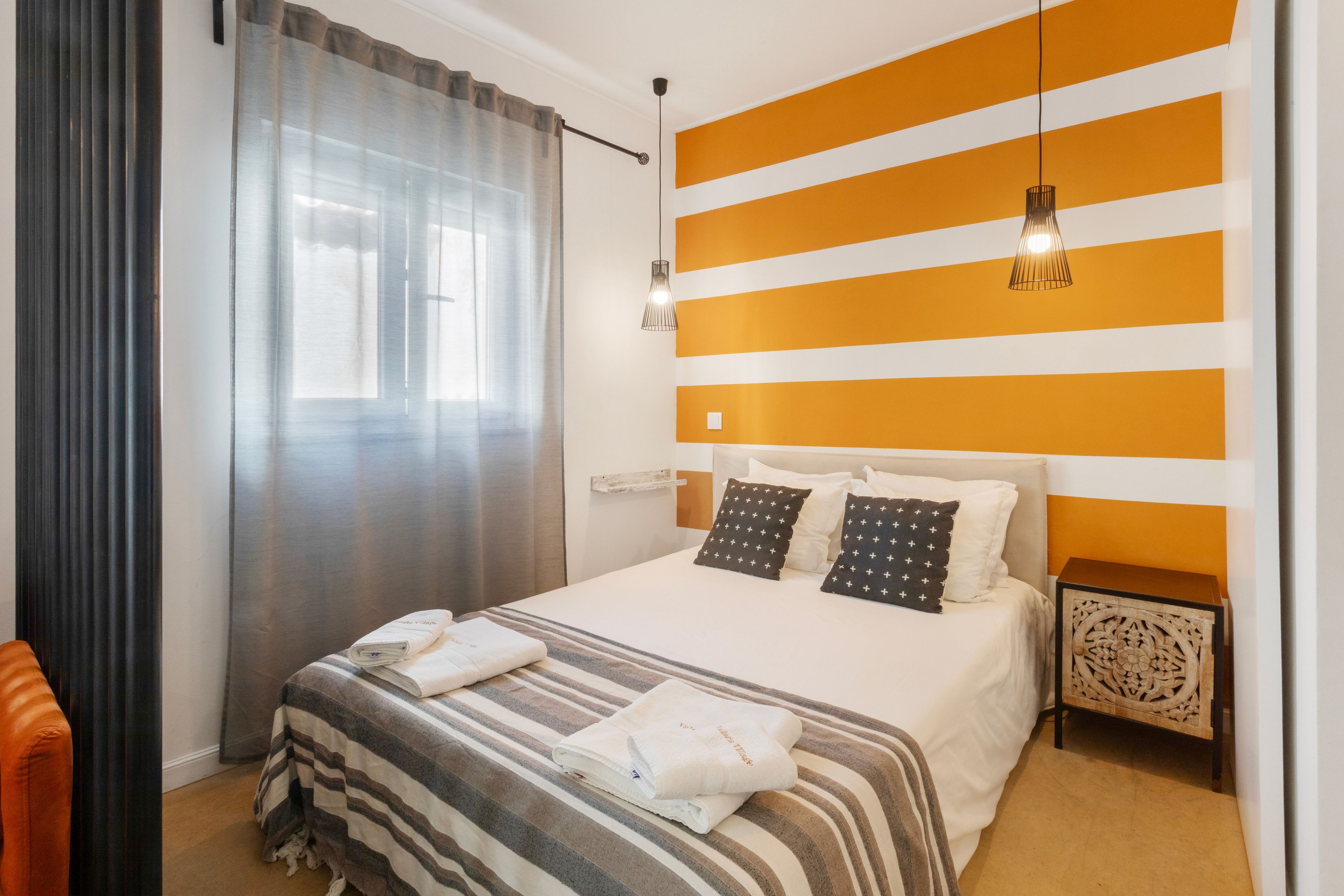 A hotel room with a bed, a nightstand, a lamp, a window with curtains, and a striped wall.
