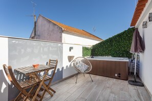 En terrasse med et træbord og stole, en fletstol, et spabad og en væg med slyngplanter.